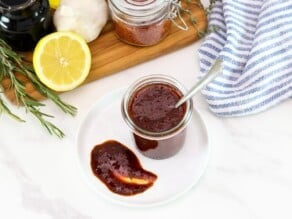 Horizontal shot of a glass jar filled with a dark red marinade sitting on top of a white plate with a dollop of sauce on the plate, fresh herbs, lemon, and pepper flakes sit in the background.