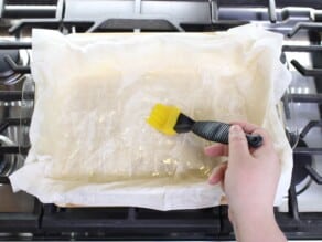 Horizontal overhead shot of a hand holding a pastry brush over a sheet of phyllo dough in a rectangular glass baking dish.