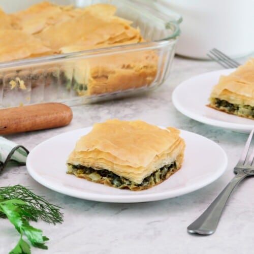 Horizontal shot of a two white plates containing square slices of vegan spanakopita. A glass dish with the remaining spanakopita is in the background to the left.