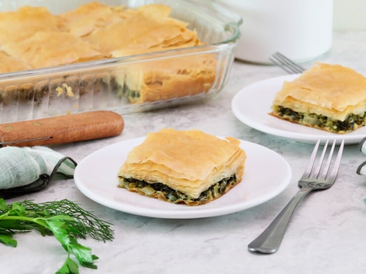 Horizontal shot of a two white plates containing square slices of vegan spanakopita. A glass dish with the remaining spanakopita is in the background to the left.