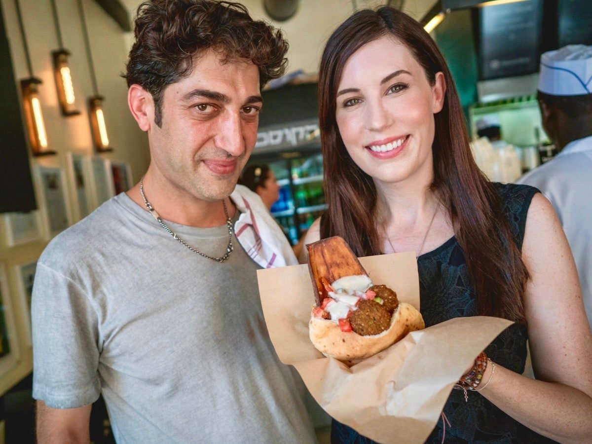 Ariel Rosenthal and Tori Avey at Hakosem restaurant in Israel. Both Ariel and Tori are smiling to the camera. Tori holds a falafel pita wrapped in paper, drizzled with tahini sauce.