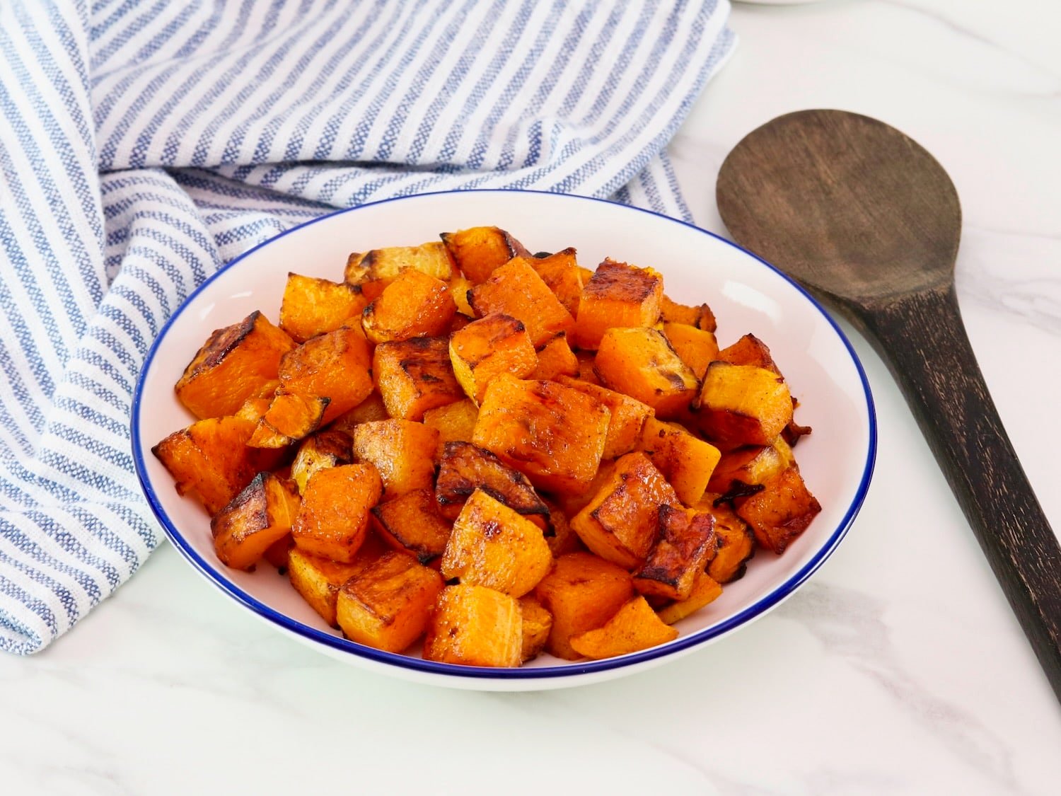Maple Roasted Butternut Squash in a large serving bowl on a marble countertop. Blue and white linen striped cloth towel and dark wooden spoon beside the bowl.