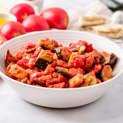 Horizontal shot - a bowl of roasted eggplant salad with red peppers in a white bowl on a white marble countertop. Fresh tomatoes, dish of olive oil peppers and pita in the background.