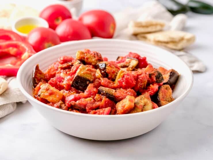 Horizontal shot - a bowl of roasted eggplant salad with red peppers in a white bowl on a white marble countertop. Fresh tomatoes, dish of olive oil peppers and pita in the background.
