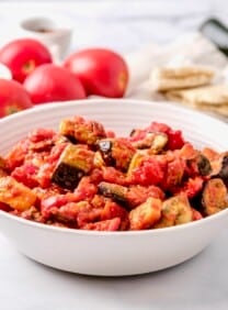Featured wide horizontal shot - a bowl of roasted eggplant salad with red peppers in a white bowl on a white marble countertop. Fresh tomatoes, dish of olive oil peppers and pita in the background.