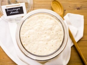 Overhead shot of large glass container on table with sourdough starter. Wooden spoon and towel laying beside it. Small sign with words handwritten "sourdough starter" near the container.