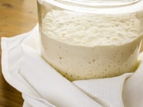 Side view of sourdough starter in a glass container with towel on wooden table.
