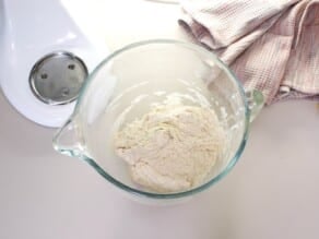 Mixed dough in glass dough ball next to stand mixer, linen towel on countertop.