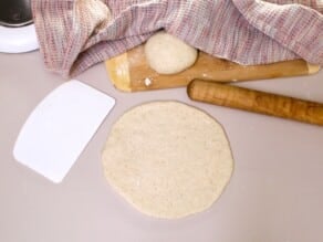 Sourdough pita dough rolled out into a circle on a smooth flat rolling surface. Dough scraper and wooden rolling pin beside it. Cutting board with a dough round covered by towel in background.