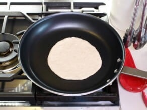 Pita dough rolled into a flat round on a nonstick skillet on a gas stovetop. Canister with tongs in background, red ceramic spoon rest next to the range.