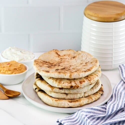 Horizontal close up - Pile of freshly baked pita breads on a plate, striped blue and white linen napkin in foreground, two small dishes of dip in background - hummus and yogurt dip - with a wooden spoon and a flour canister in the background, as well.