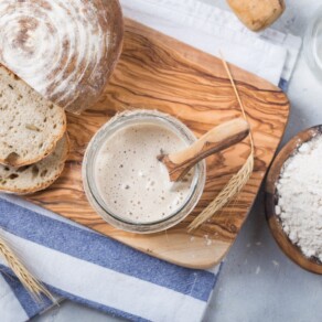 Overhead shot of sourdough starter with wooden spoon in glass bowl on a wooden cutting board. Sourdough boule sliced alongside the bowl. Dish of dry flour beside the cutting board. Scene is decorated by shaft of wheat.