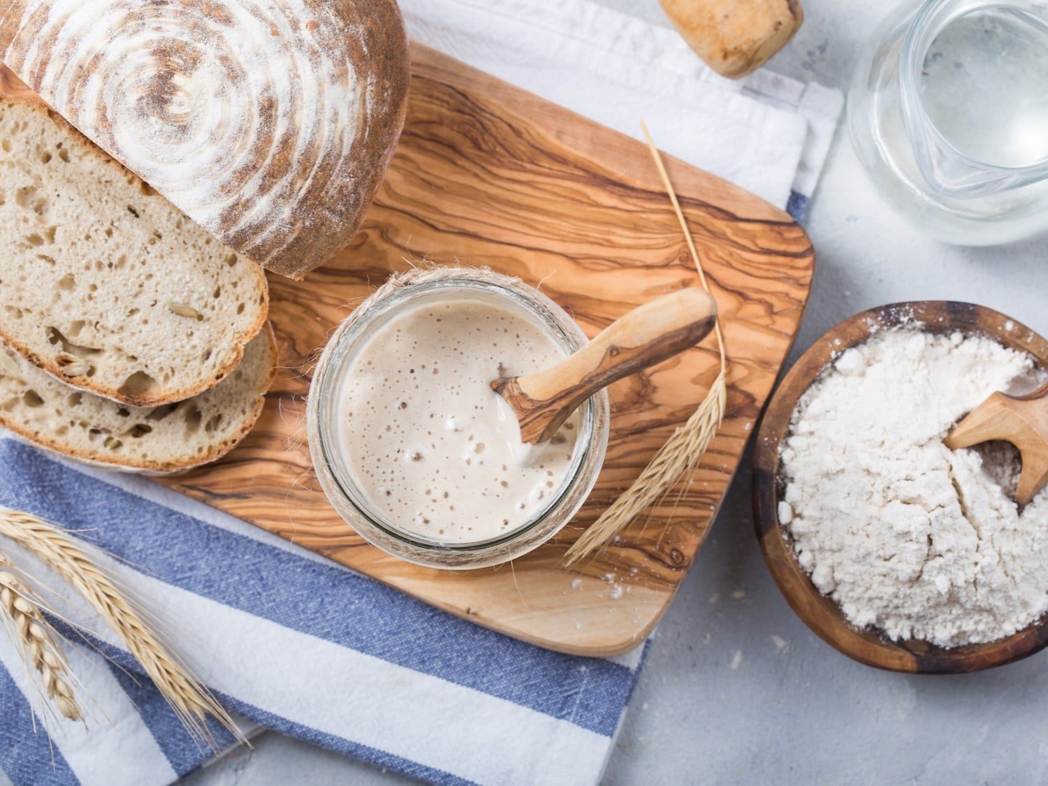 Overhead shot of sourdough starter with wooden spoon in glass bowl on a wooden cutting board. Sourdough boule sliced alongside the bowl. Dish of dry flour beside the cutting board. Scene is decorated by shaft of wheat.