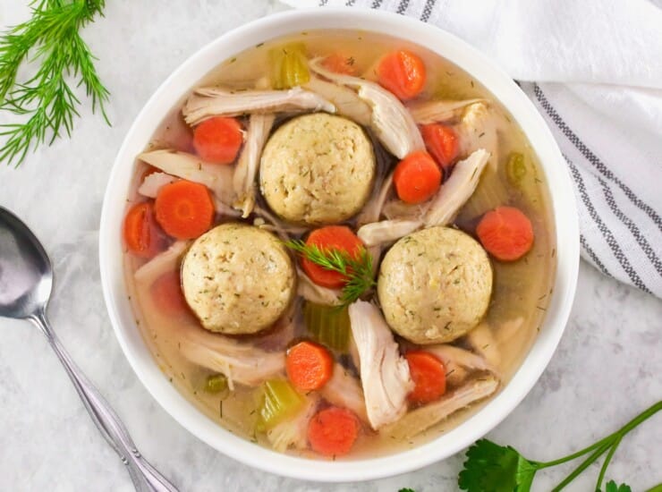 Overhead shot of a bowl of floater matzo balls in a shallow bowl of Jewish chicken soup with carrot slices, pieces of celery, and golden broth. Spoon, fresh herbs, and linen napkin on the white marble counter beside the bowl.