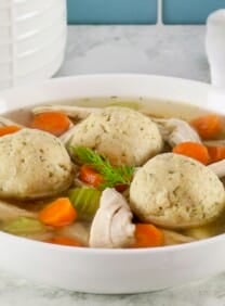 Horizontal shot of floater matzo balls in a shallow bowl of Jewish chicken soup with carrot slices, pieces of celery, and golden broth. Spoon and linen napkin on the white marble counter beside the bowl. Tiles and white jar in background.