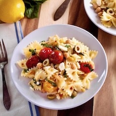Plate of lemon pasta salad with roasted tomatoes, chickpeas, olives and feta on a wooden cutting board with linen napkin and fork. Lemon and second plate of salad in background.