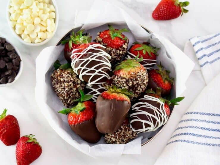 Chocolate dipped strawberries, some with nuts and some chocolate strawberries drizzled with white chocolate on a parchment lining paper inside a medium-size circular gold tin can in foreground, with a linen napkin on the side. Three fresh strawberries, and two small bowls with white and dark chocolate chips separately in background.