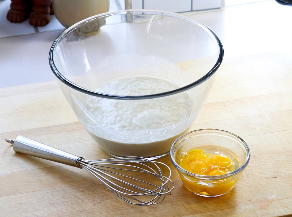 A glass bowl with batter and a smaller bowl with cracked eggs sit on a wooden counter, accompanied by a metal whisk.