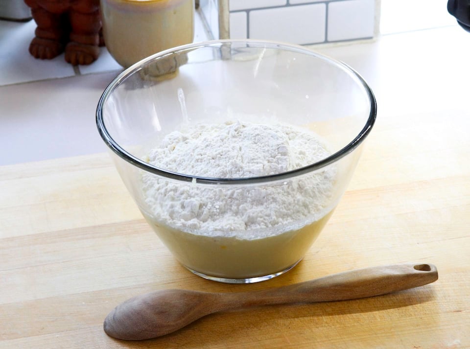 A glass bowl filled with flour rests on a wooden surface, beside a wooden spoon.