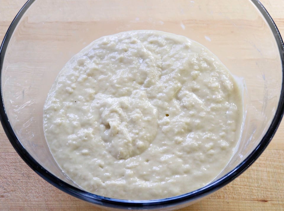 A clear glass bowl filled with yeast starter sits on a wooden surface.