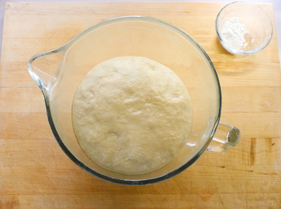 Dough rising in a transparent glass bowl on a wooden surface, with a small bowl of flour nearby.