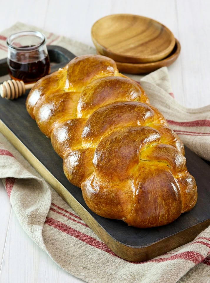 Golden braided challah bread with a jar of honey and dipper beside it, placed on a dark wooden chopping board with a rustic red-striped kitchen towel underneath. Wooden small plates in the background.