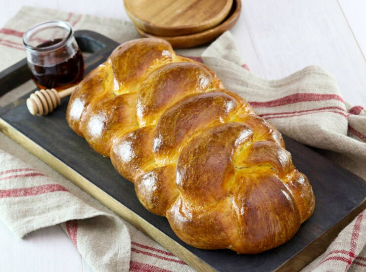 Golden braided challah bread with a jar of honey and dipper beside it, placed on a dark wooden chopping board with a rustic red-striped kitchen towel underneath. Nearby are wooden plates.