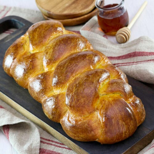 Golden braided challah bread placed on a dark wooden chopping board with a rustic red-striped kitchen towel underneath. Nearby are a jar of honey, honey dipper and wooden plates.