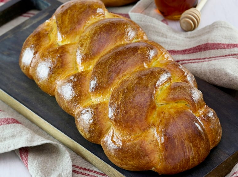 Golden braided challah bread placed on a dark wooden chopping board with a rustic red-striped kitchen towel underneath. Nearby are a jar of honey and honey dipper.