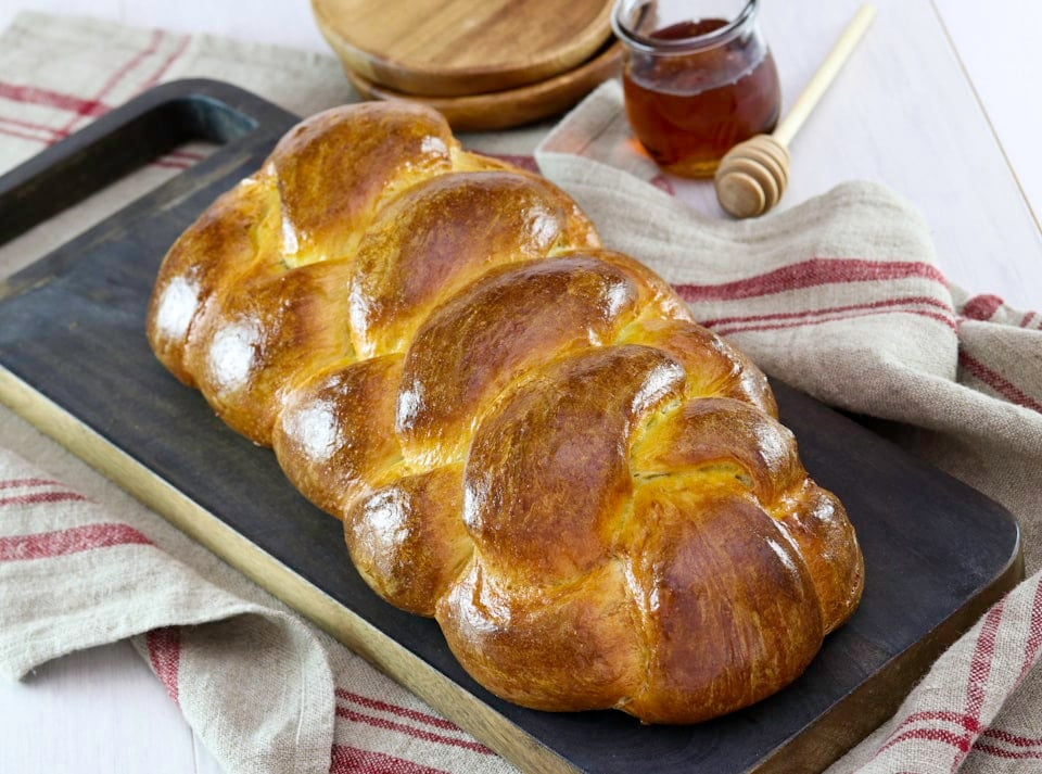 Golden braided challah bread placed on a dark wooden chopping board with a rustic red-striped kitchen towel underneath. Nearby are a jar of honey, honey dipper and wooden plates.