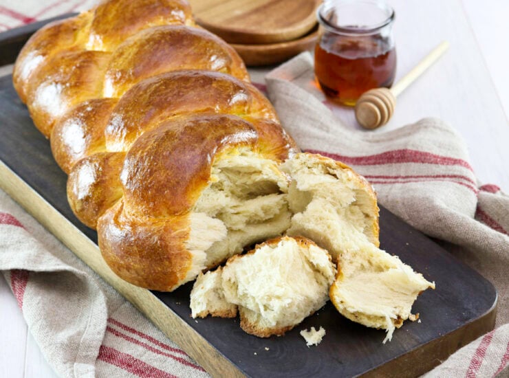 Golden braided bread sits on a dark board, partially torn to reveal its fluffy inside. Nearby are a jar of honey, a honey dipper and wooden plates.