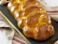 A Pinterest Sourdough Challah Pin - Golden braided sourdough challah on a dark tray, accompanied by a glass of honey. Text reads “Sourdough Challah.”.