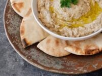 A Pinterest picture of a white bowl of baba ganoush drizzled with olive oil and garnished with parsley sits on a rustic platter, surrounded by slices of pita bread.