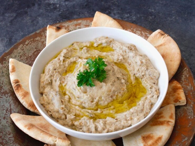 A white bowl of baba ganoush drizzled with olive oil and garnished with parsley sits on a rustic platter, surrounded by slices of pita bread.