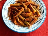 Bamya - Middle Eastern Okra Pinterest Pin. A dish of okra in tomato sauce, with spoon and small dish of chili flakes above it, on a red tablecloth.