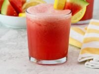 Frozen Watermelon Lemonade Pinterest Pin - close up vertical shot - frozen watermelon lemonade drinks on a countertop garnished with slices of watermelon on the glasses. On the side, a bowl of fresh watermelon slices, a striped yellow towel.