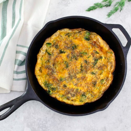 Broccoli Cheddar Frittata with greens and cheese in a black skillet on a light countertop, accompanied by a striped kitchen towel.
