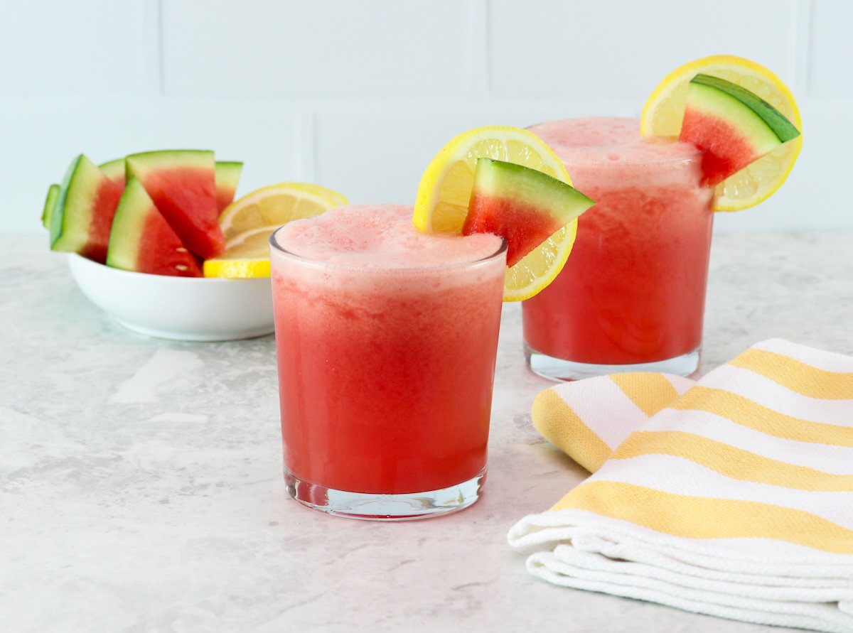 Horizontal shot - frozen watermelon lemonade drinks on a countertop garnished with slices of watermelon on the glasses. On the side, a bowl of fresh watermelon slices, a striped yellow towel.
