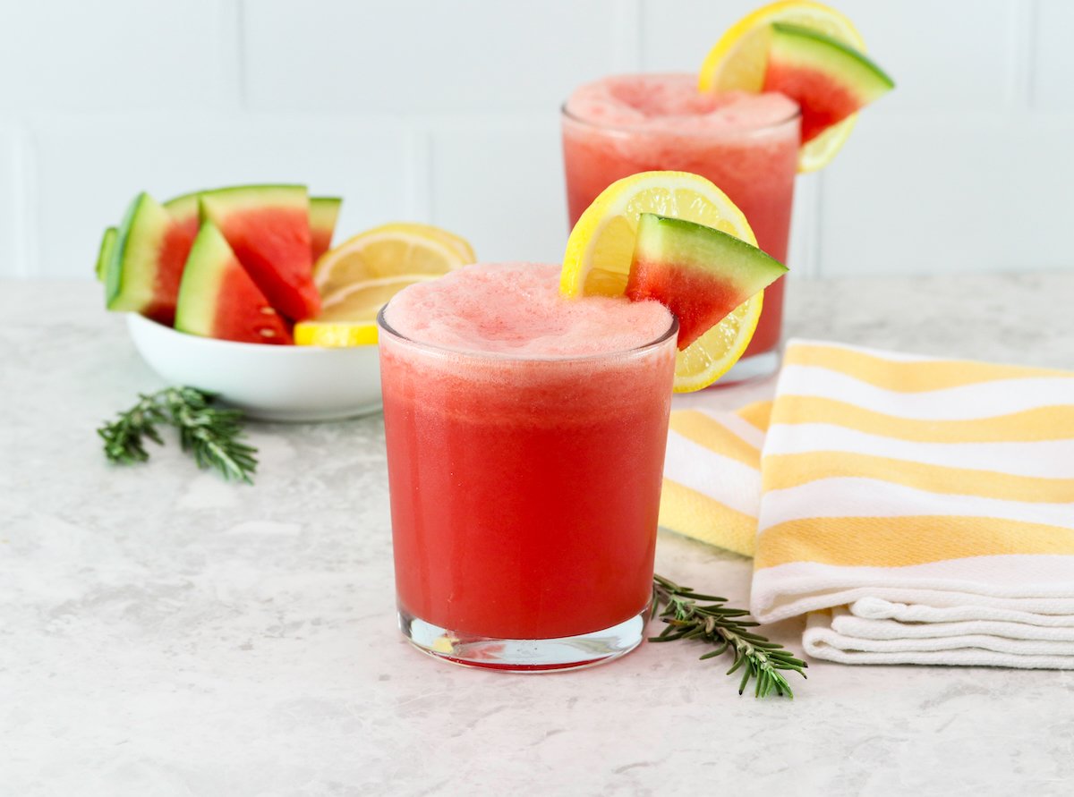 Two frozen watermelon lemonade drinks on a countertop garnished with slices of watermelon on the glasses. On the side, a bowl of fresh watermelon slices, a striped yellow towel, and some fresh rosemary sprigs.
