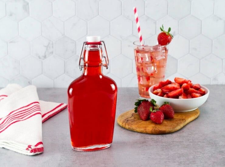 A bottle of Homemade Strawberry Syrup next to a tall glass of pink iced strawberry drink with a straw and a red-striped towel. Fresh strawberries and a bowl of sliced strawberries are on a cutting board.