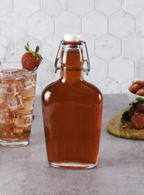 A bottle of Homemade Strawberry Syrup next to a tall glass of pink iced strawberry drink with a straw and a red-striped towel. Fresh strawberries and a bowl of sliced strawberries are on a cutting board.