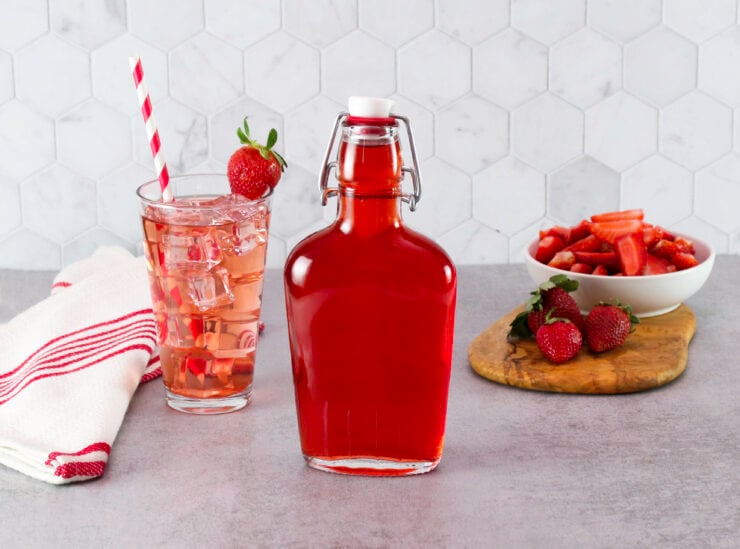 A bottle of Homemade Strawberry Syrup next to a tall glass of pink iced strawberry drink with a straw and a red-striped towel. Fresh strawberries and a bowl of sliced strawberries are on a cutting board.