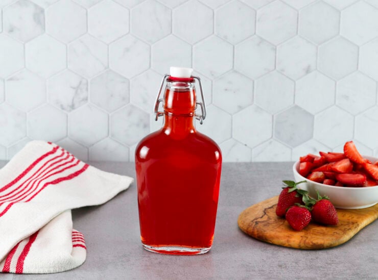 A bottle of Homemade Strawberry Syrup next to a red-striped towel. Fresh strawberries and a bowl of sliced strawberries are on a cutting board.