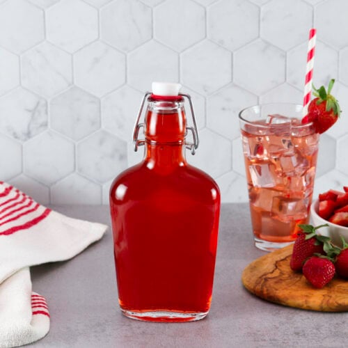 A bottle of Homemade Strawberry Syrup next to a tall glass of pink iced strawberry drink with a straw and a red-striped towel. Fresh strawberries and a bowl of sliced strawberries are on a cutting board.
