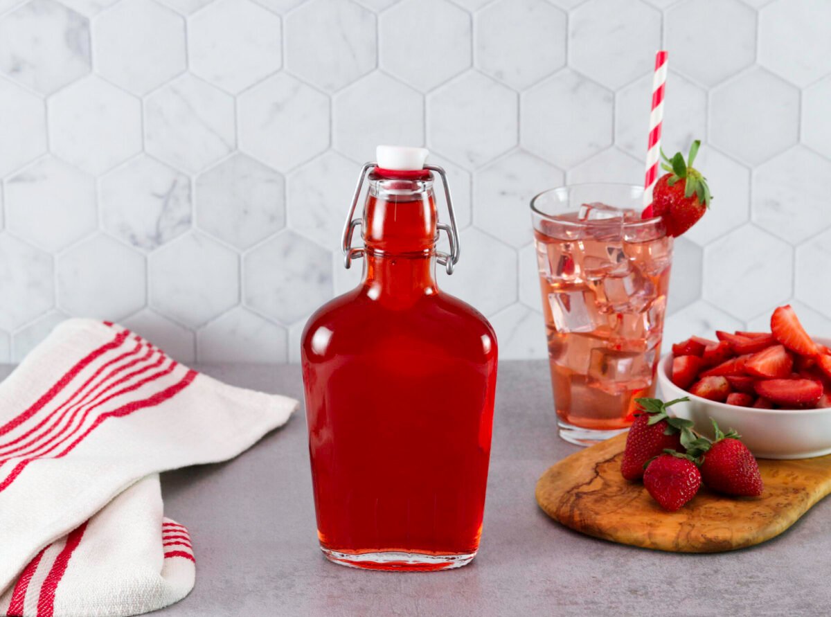 A bottle of Homemade Strawberry Syrup next to a tall glass of pink iced strawberry drink with a straw and a red-striped towel. Fresh strawberries and a bowl of sliced strawberries are on a cutting board.