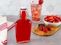 Simple Strawberry Syrup Recipe Pinterest Pin - bottle of bright red strawberry syrup with a white stopper on a concrete countertop, red and white cloth towel on the side, wooden cutting board with fresh strawberries, whole and sliced, and a glass of strawberry soda with a straw, garnished with a fresh strawberry, in the background.