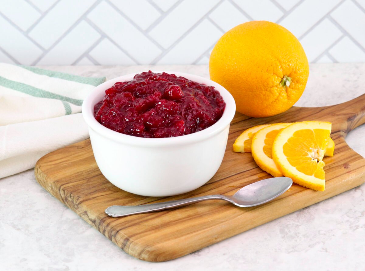 A bowl of cranberry sauce on a wooden board, surrounded by sliced and whole oranges, a spoon and a towel is draped beside it.