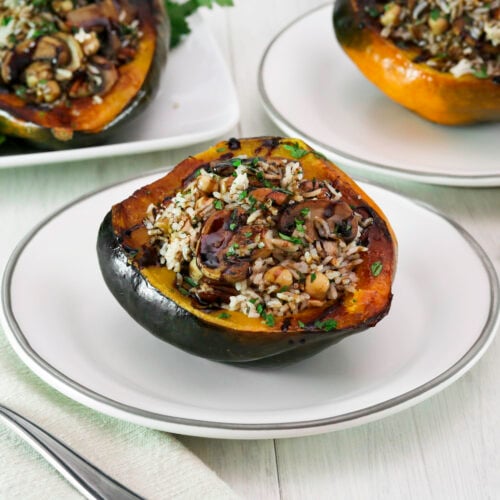 Vegan stuffed acorn squash is filled with rice, mushrooms, chickpeas, and herbs on a white plate. A fork is placed on a beige napkin beside it.