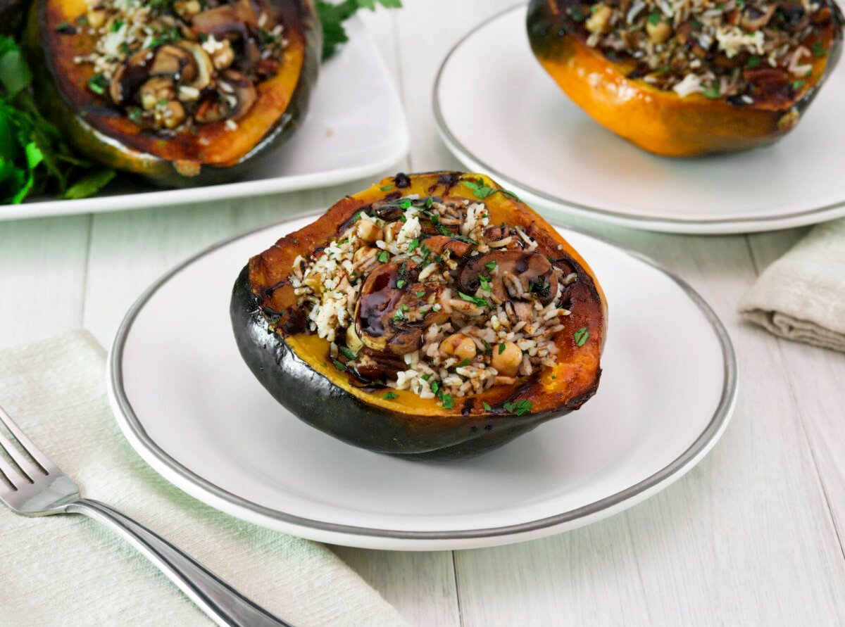 Vegan stuffed acorn squash is filled with rice, mushrooms, chickpeas, and herbs on a white plate. A fork is placed on a beige napkin beside it.