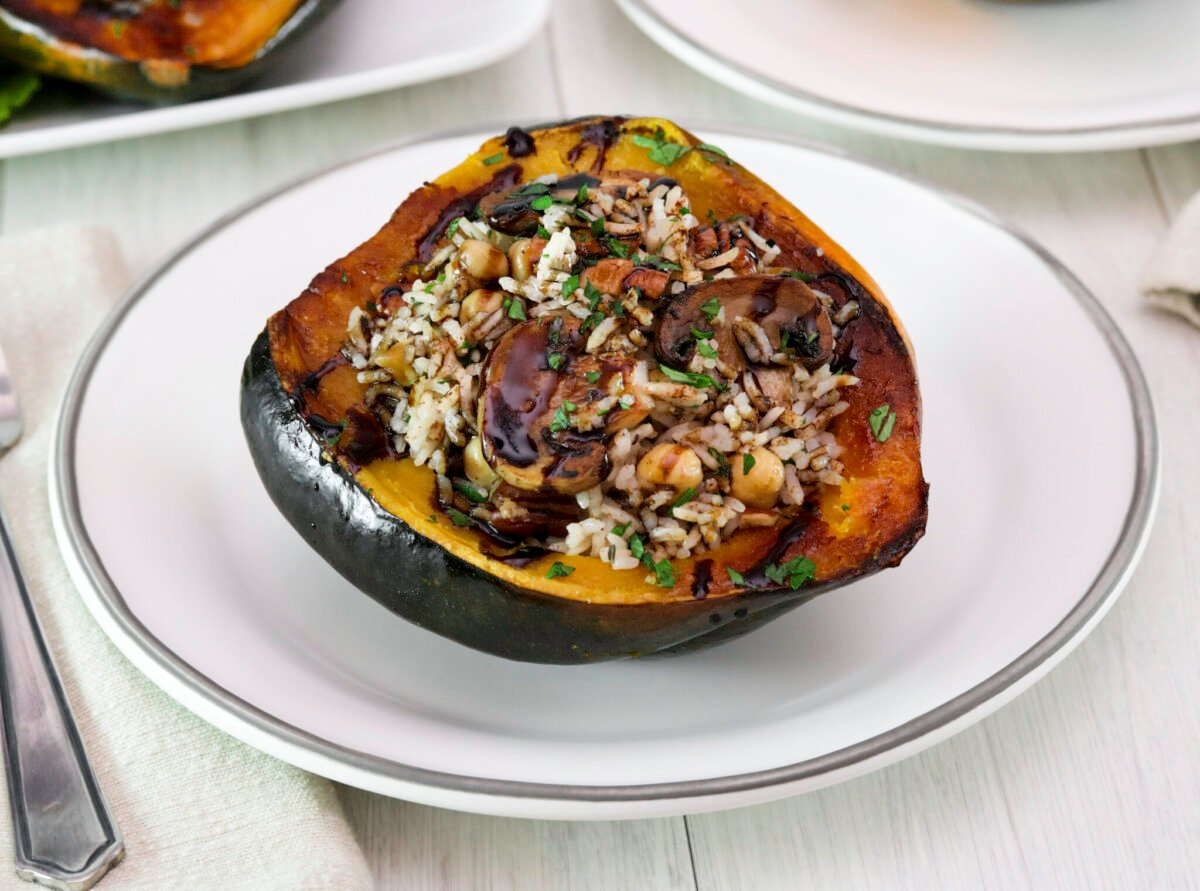 Vegan stuffed acorn squash filled with wild rice, mushrooms, and herbs on a white plate. Light wood table, and white tile background. A fork is placed on a beige napkin beside it.